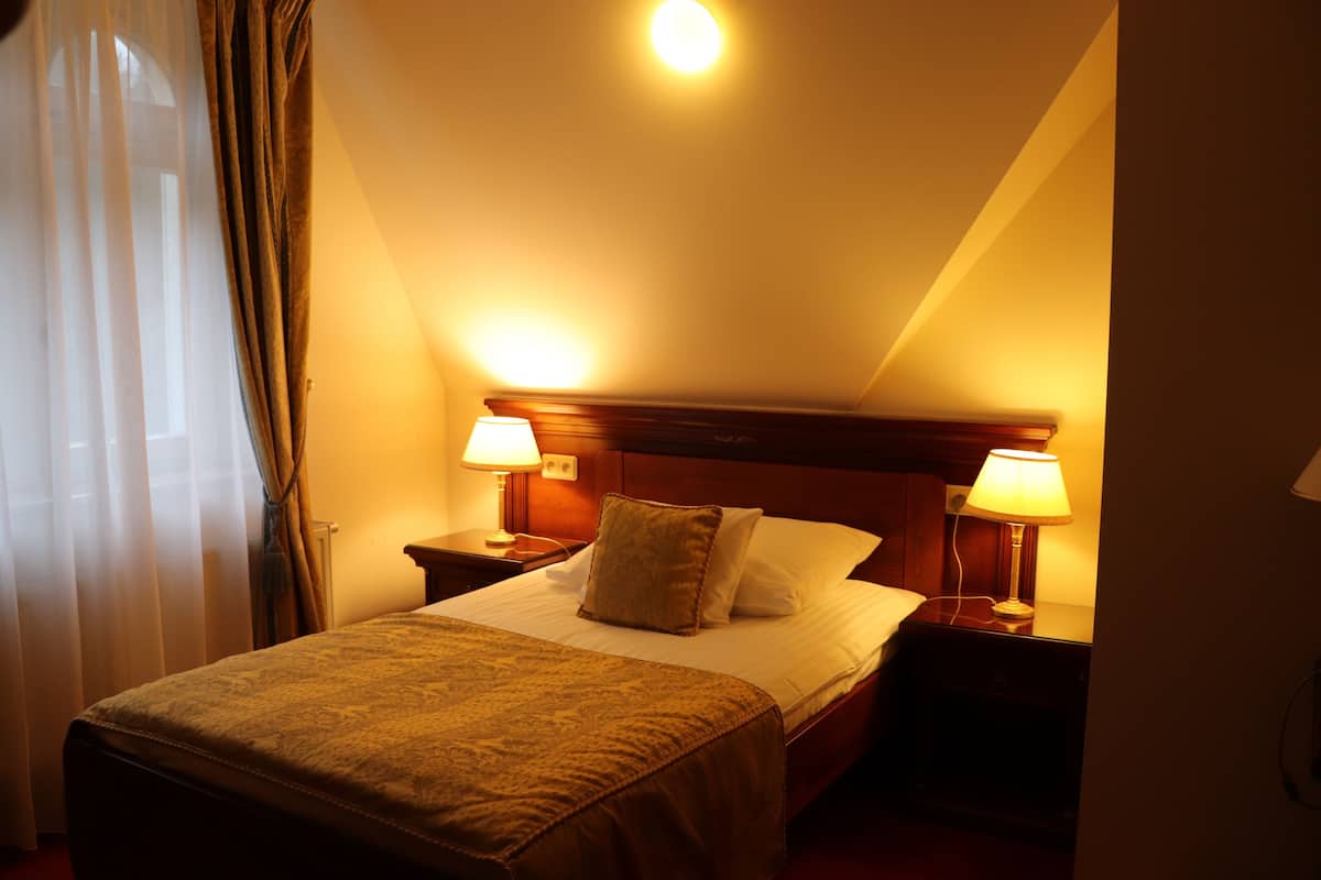 Quarto de hotel com cama, duas luminárias de cabeceira, mesa de apoio e janela com cortinas.