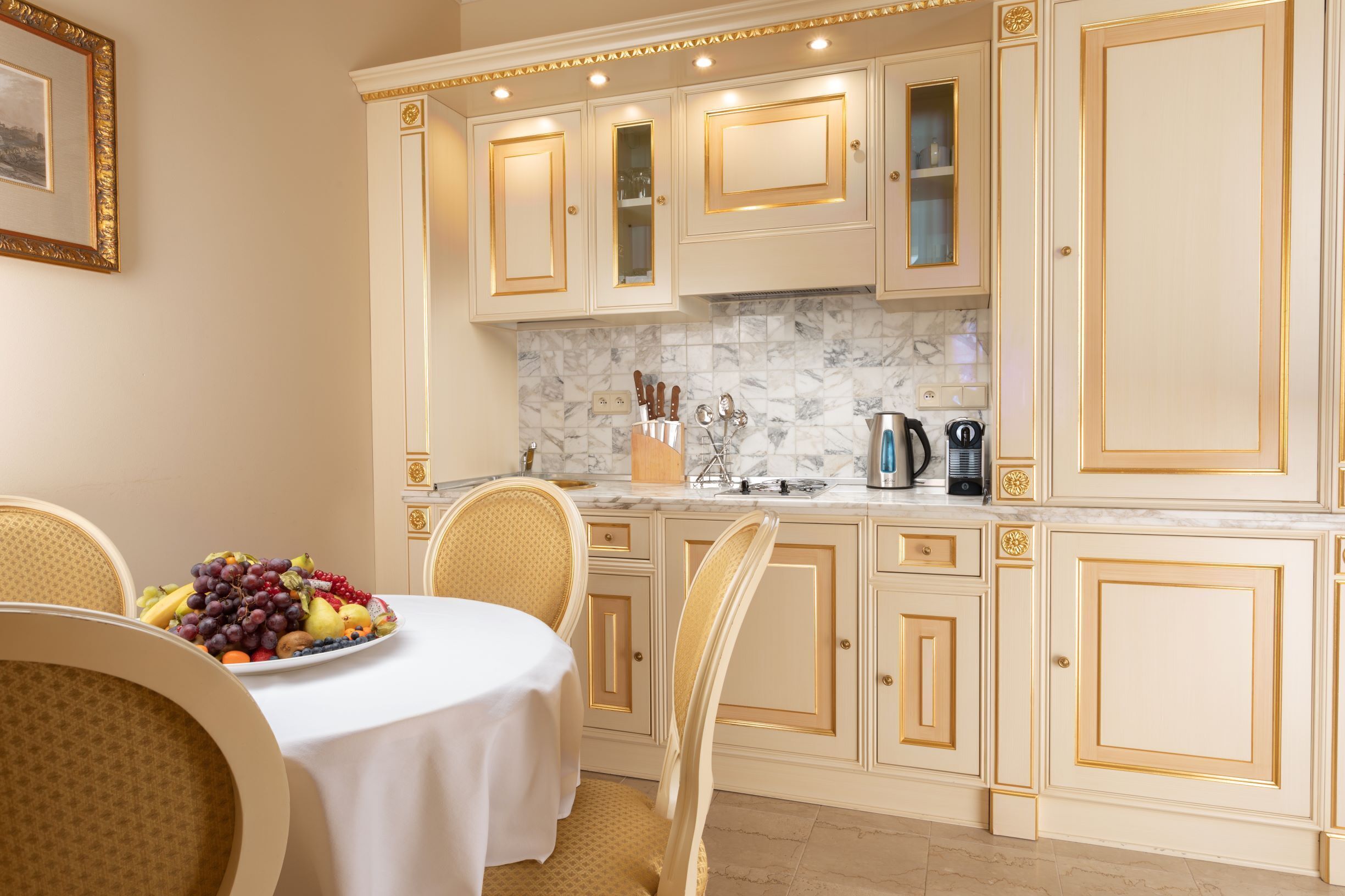 A kitchen with white cabinetry and marble backsplash, a dining table with a fruit platter, and a coffee maker.