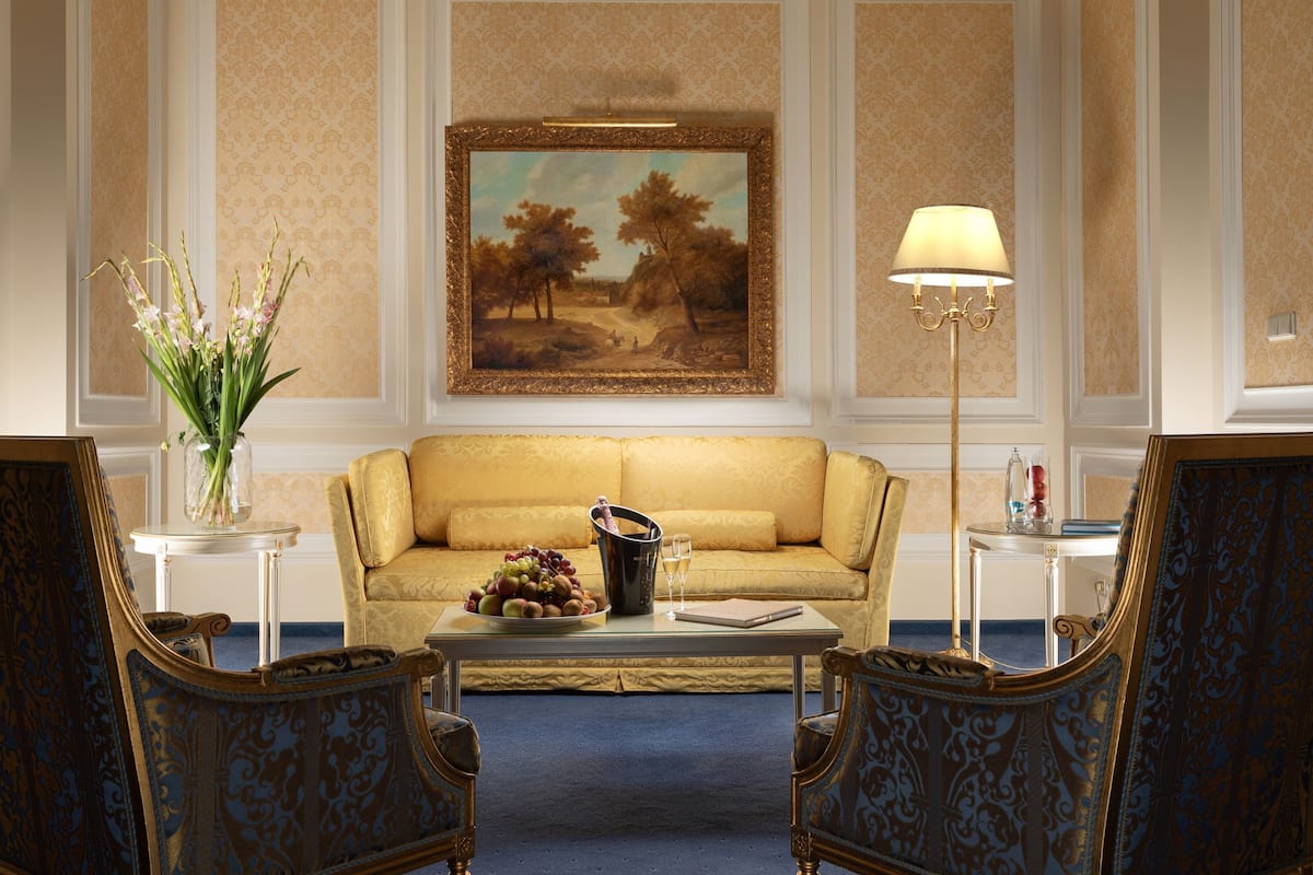 Um quarto com um sofá, duas poltronas, uma mesa pequena com um vaso de flores, um quadro emoldurado na parede e um abajur.