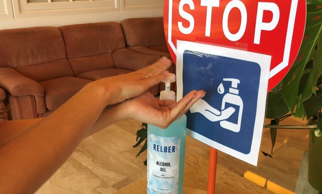 Une main distribue du gel hydroalcoolique à partir d’un flacon étiqueté « Gel alcoolisé RELBER », à côté d’un panneau de signalisation représentant un symbole de lavage des mains.