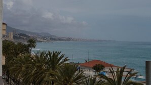 A coastal view with a building featuring a red roof, palm trees, and a view of the sea and distant cityscape.