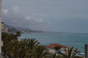 A coastal view with a building featuring a red roof, palm trees, and a view of the sea and distant cityscape.