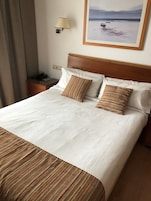 Habitación de hotel con una cama grande, dos almohadas, una mesita de noche, una lámpara y un cuadro de una playa.