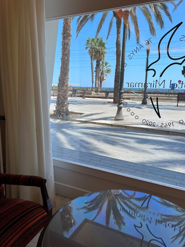 A view from a window with a table and chair inside, overlooking a street with palm trees and a blue sky.
