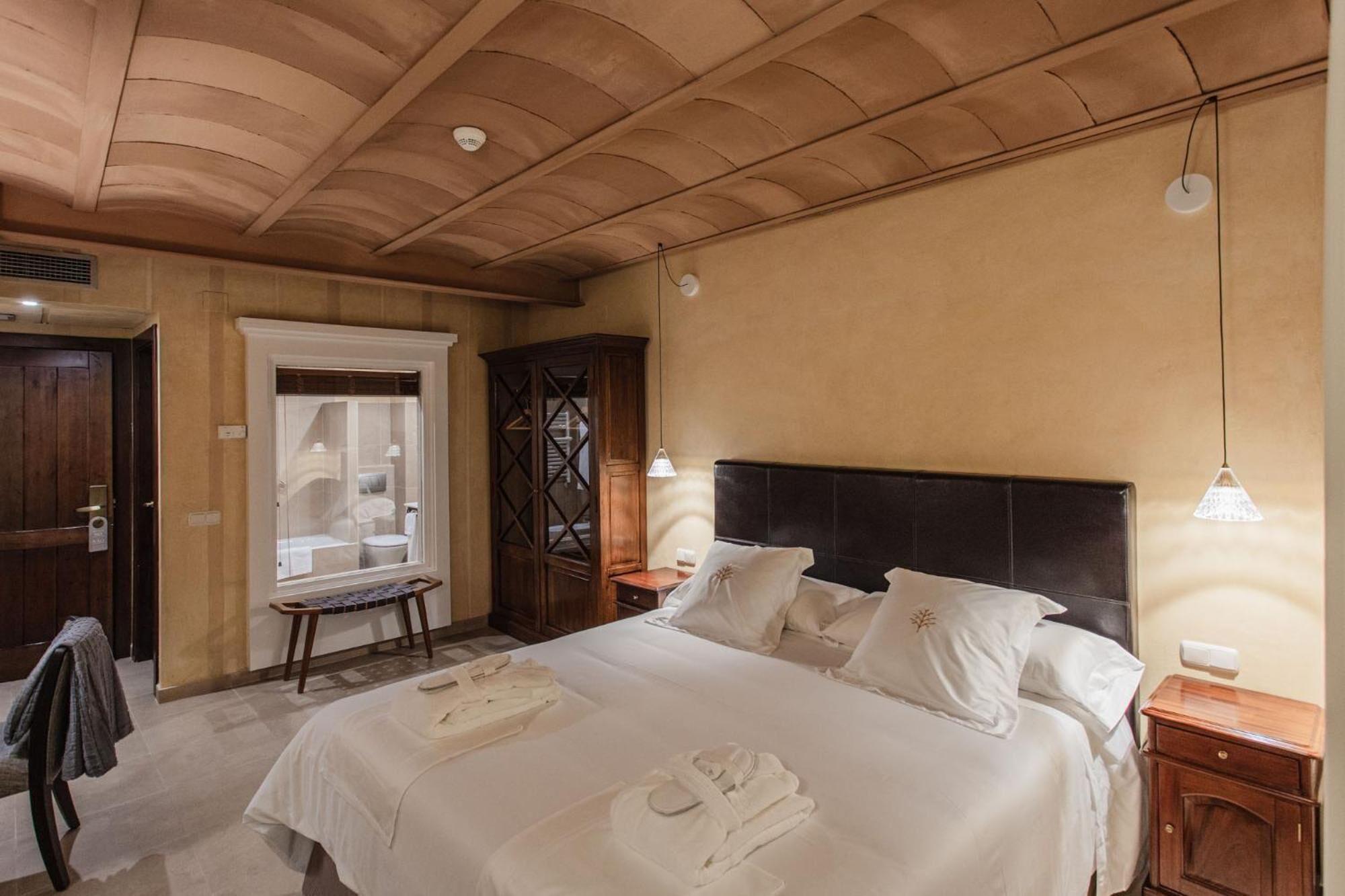 A hotel room with a large bed, a wooden wardrobe, a small table, and a chair.