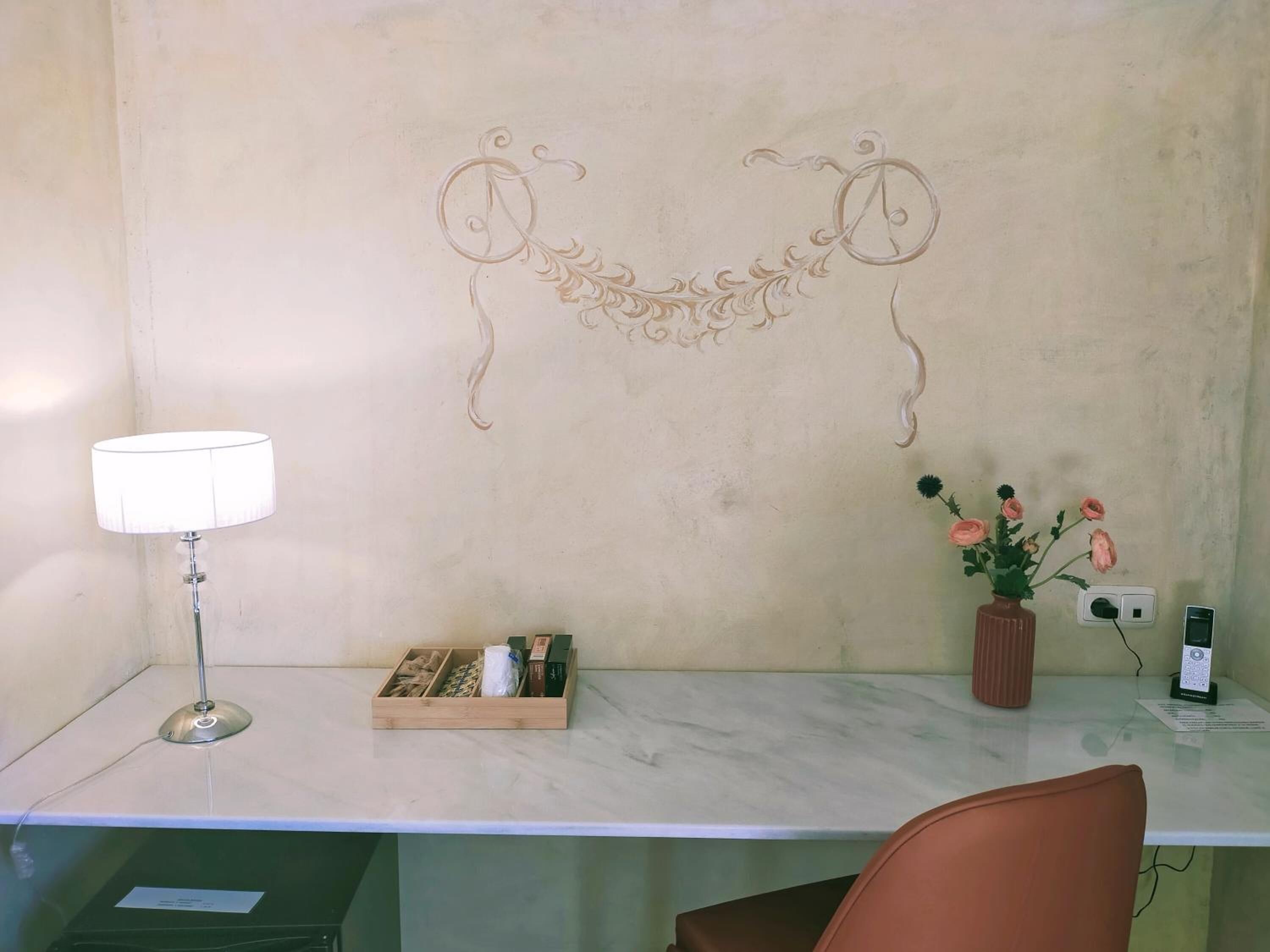 Un escritorio con una lámpara, un jarrón con flores, una caja con objetos y un diseño decorativo en la pared.