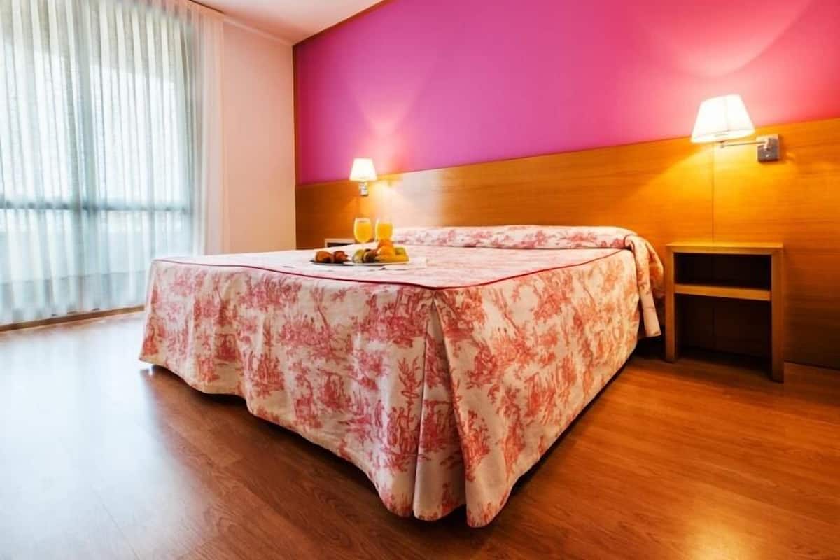 Habitación de hotel con cama, mesita de noche con lámpara, ventana con cortinas y pared con detalle en rosa.
