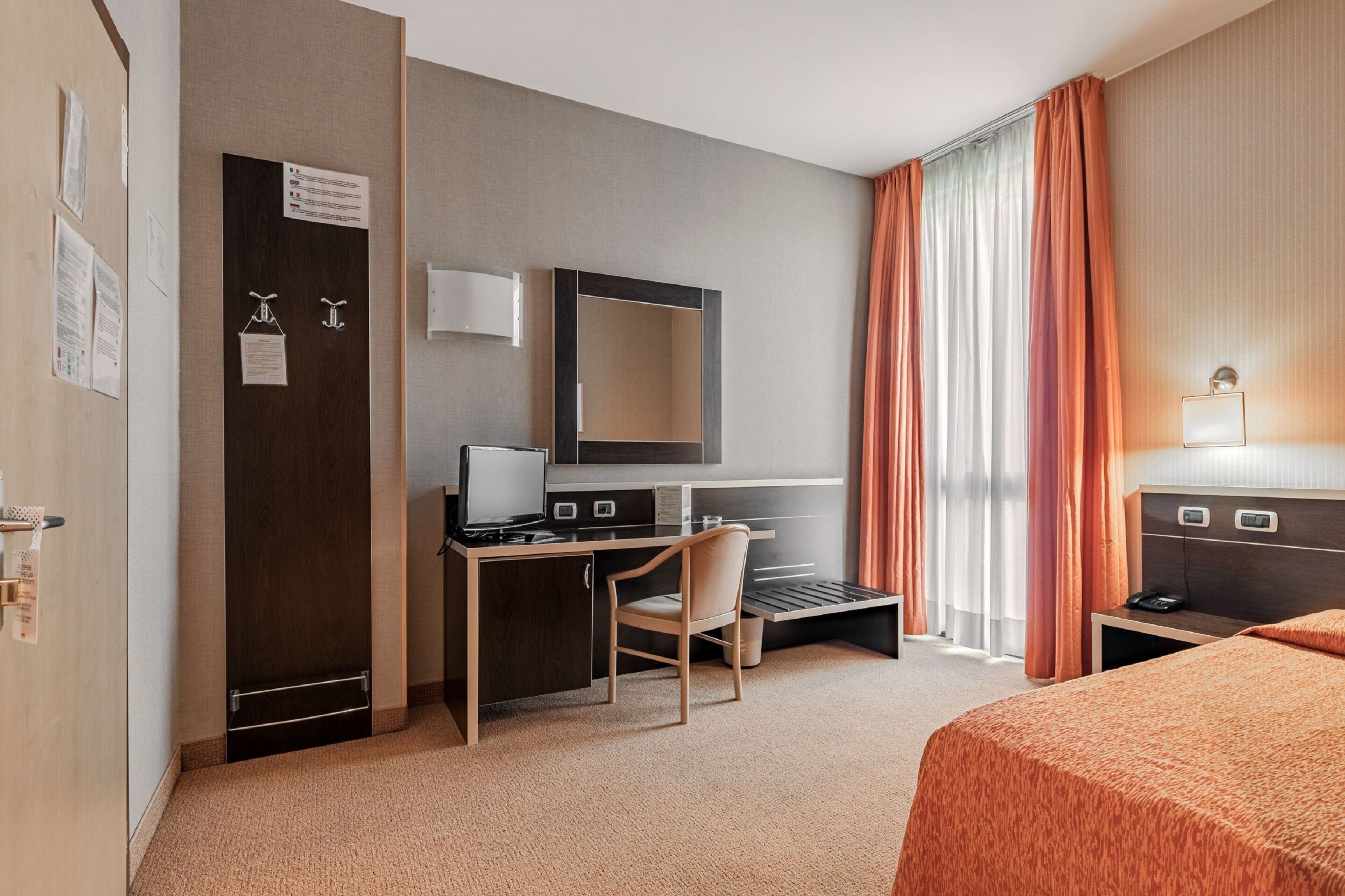 A hotel room with a desk, chair, bed, and a large window with curtains.