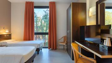 Habitación de hotel con dos camas, un escritorio con televisión y vistas a árboles por la ventana.