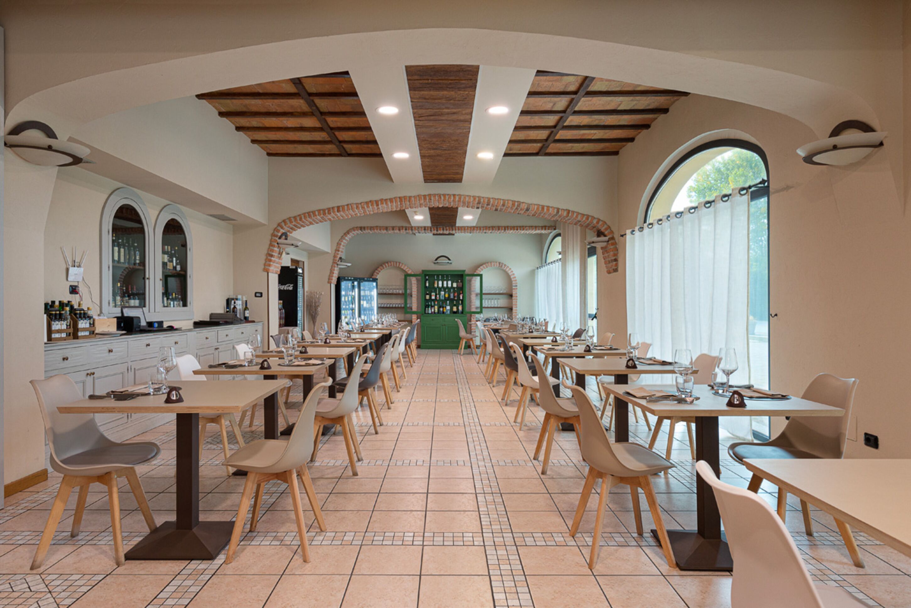 A spacious restaurant with high ceilings, arched brick arches, and a central green counter. Tables are arranged in rows with chairs.