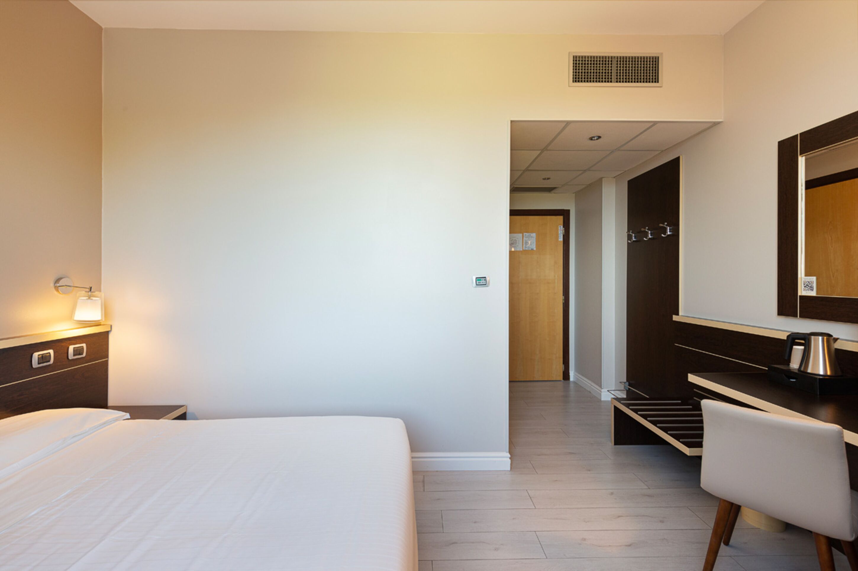 Una habitación de hotel con cama, mesita de noche, lámpara, escritorio con silla y espejo.