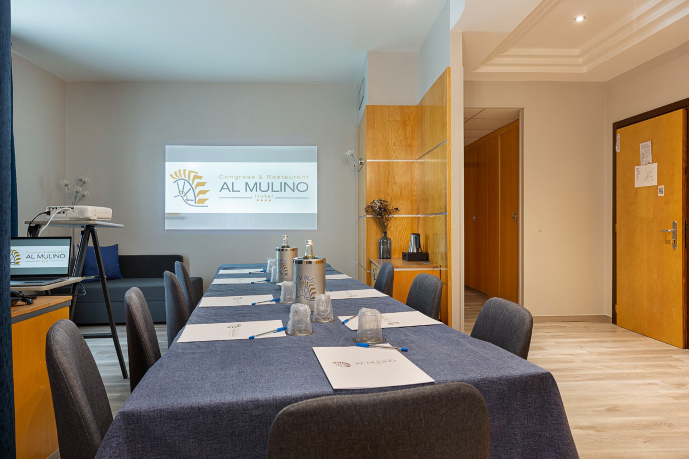 A conference room with a long table, chairs, a projector screen displaying the Al Mulino logo, a laptop, and a coffee machine.