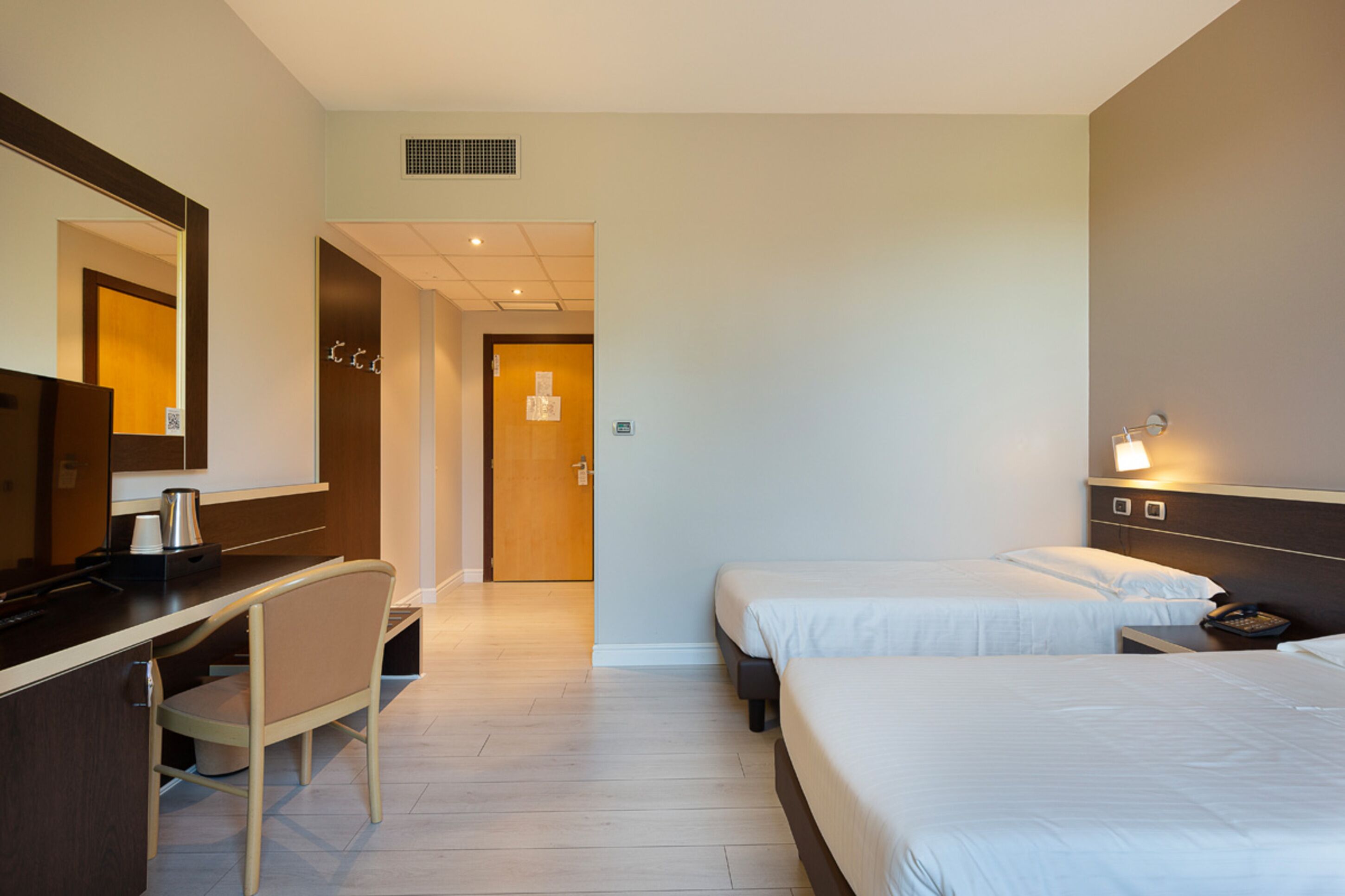 A hotel room with a desk, two beds, a chair, and a mirror.
