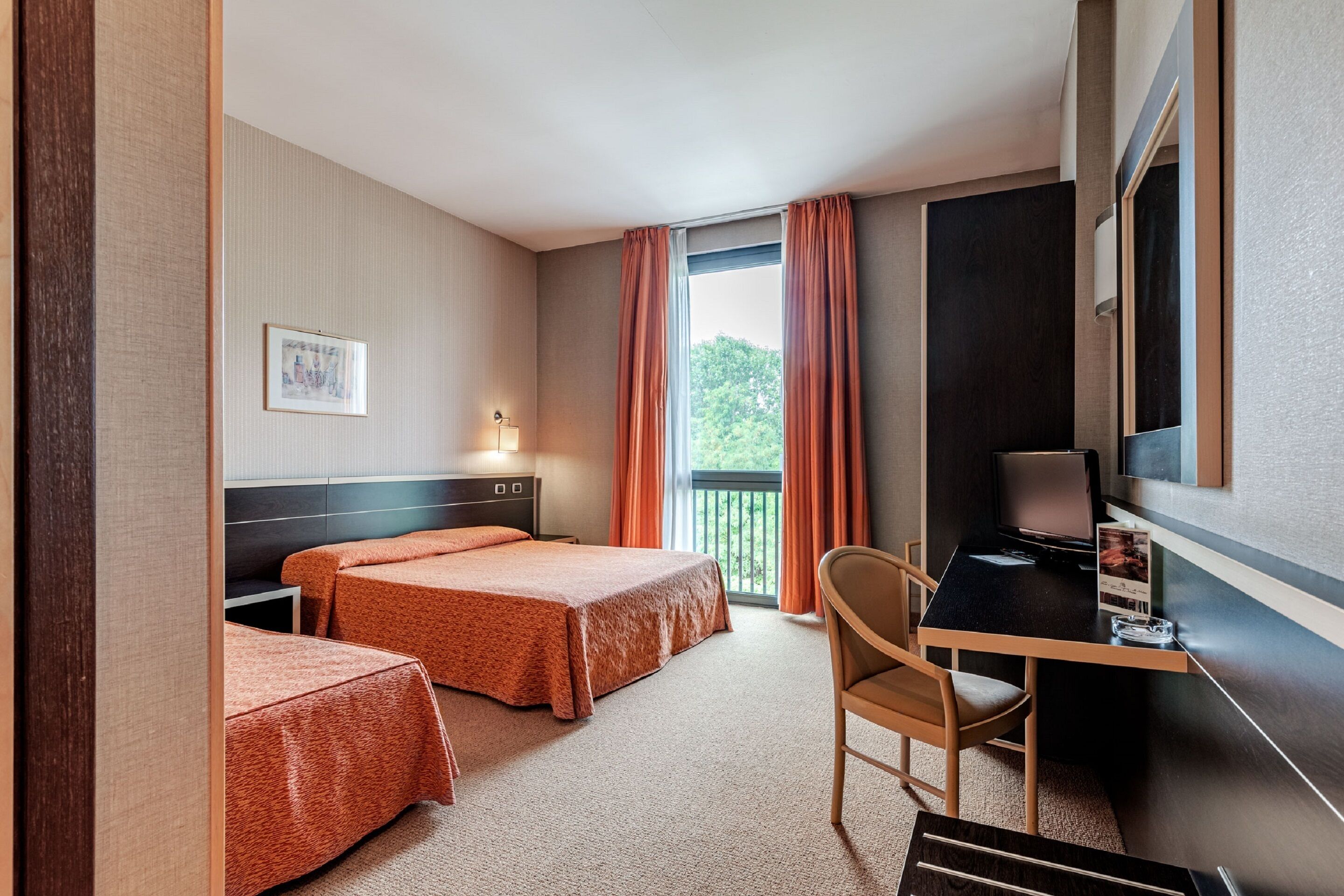 Habitación de hotel con dos camas, un escritorio, una silla, un televisor y un balcón con cortinas.