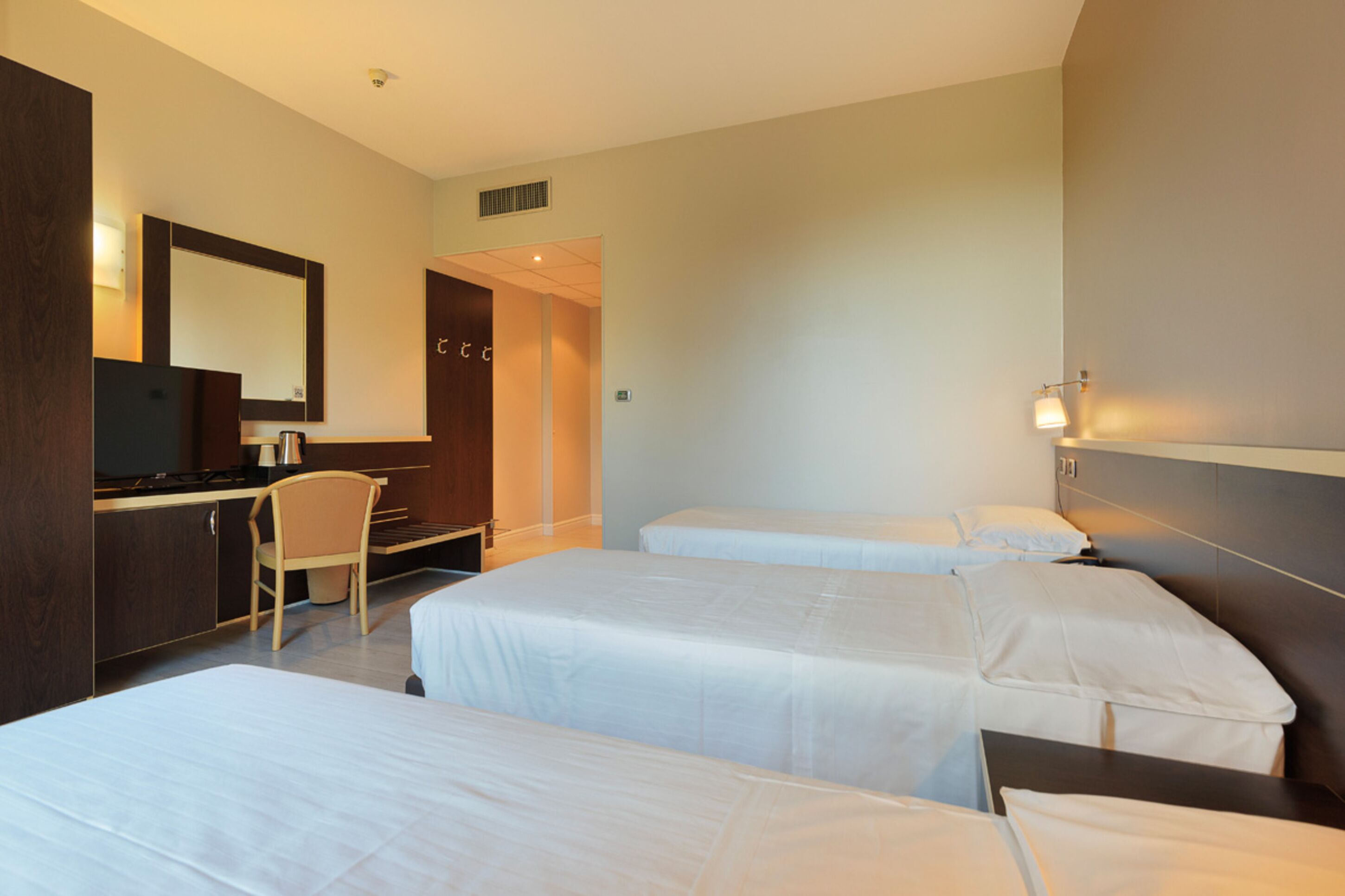 A hotel room with two single beds, a desk with a chair, a mirror, and a small kitchenette.