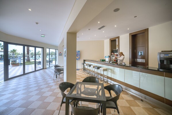 Un área de bar moderna con una mesa y sillas de vidrio, una barra con taburetes y vistas al patio exterior con palmeras.