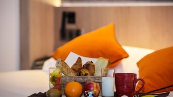 Ein Frühstückswagen mit Früchten, Gebäck und Getränken auf einem Bett mit orangefarbenen Kissen.
