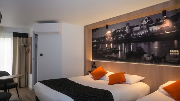 A hotel room with a large bed, two orange pillows, a black throw blanket, and a wall-mounted painting of a nighttime cityscape.