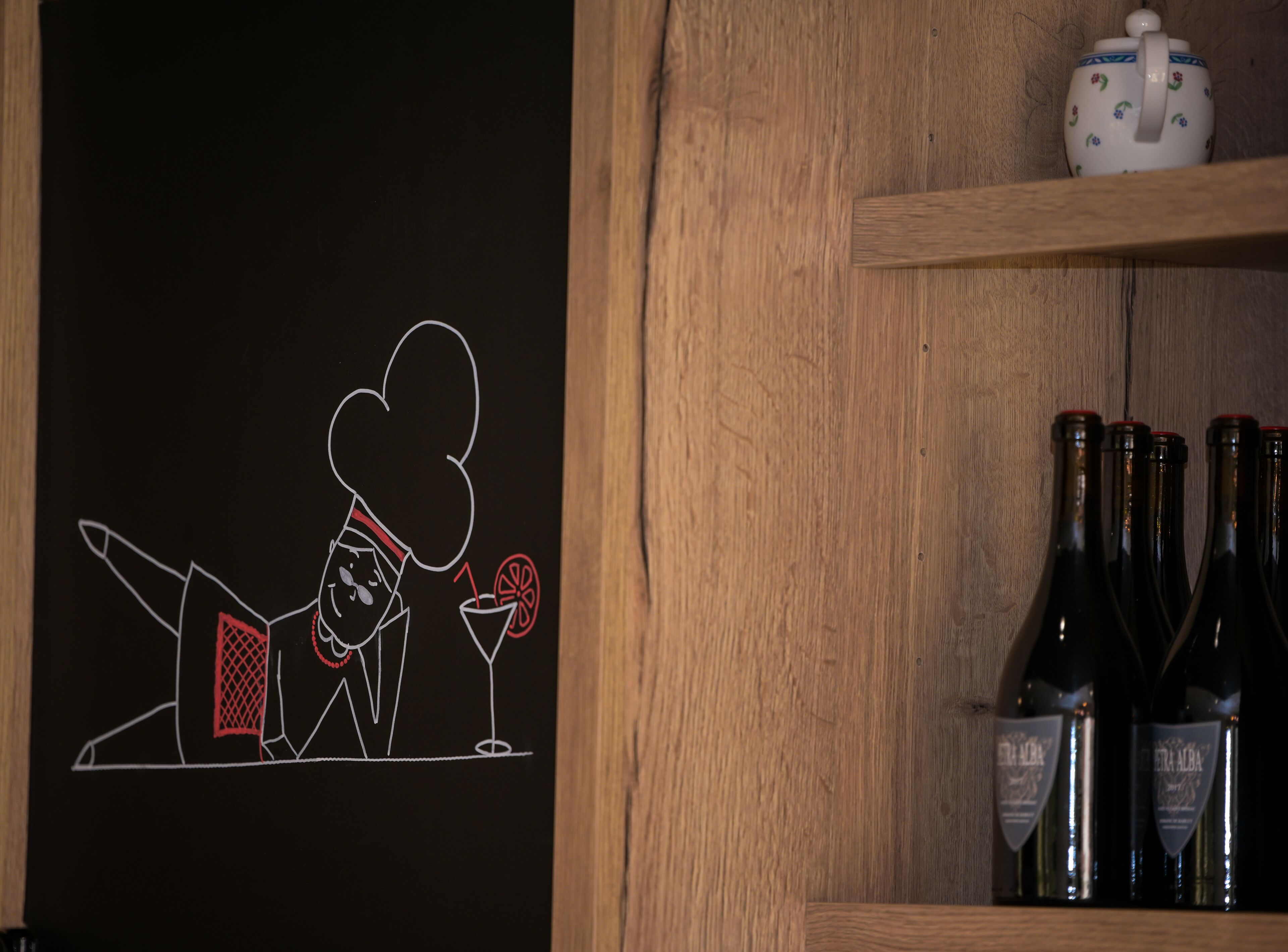 A chalkboard with a chef illustration, a teapot, and wine bottles on shelves.