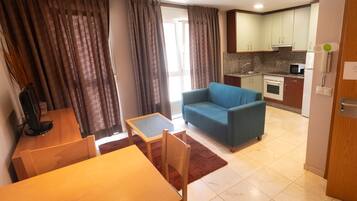 A compact living space with a dining area, a sofa, a kitchen with basic appliances, and a view through a window with curtains.