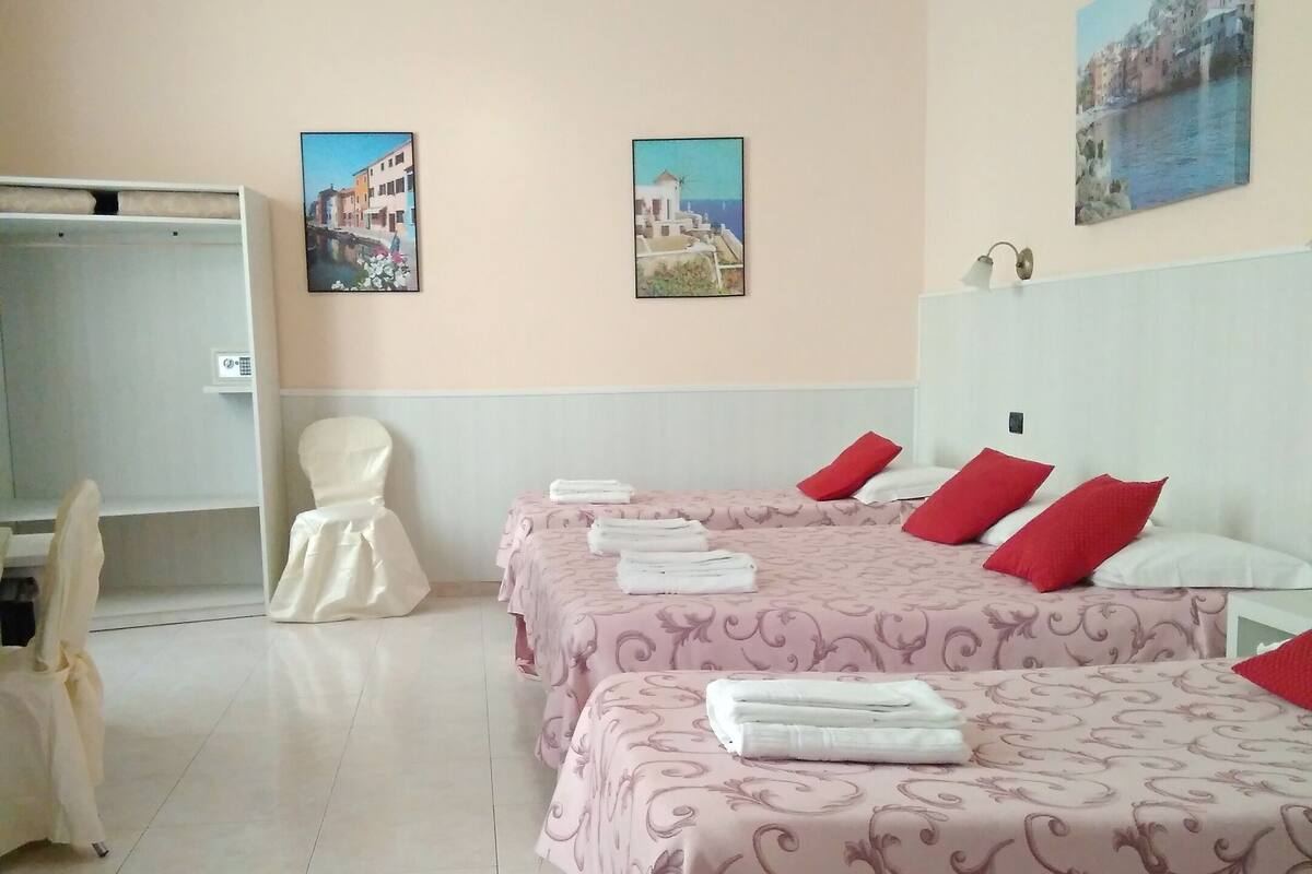 A hotel room with two beds, each with pink patterned bedsheets and red pillows, a white headboard, and a ceiling fan. There are two wall-mounted paintings depicting coastal scenes.