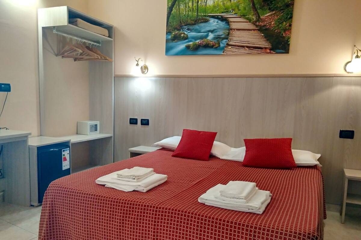 A hotel room with a bed, red bedding, two pillows, a painting of a forest stream, a desk with a computer, and a small cabinet.