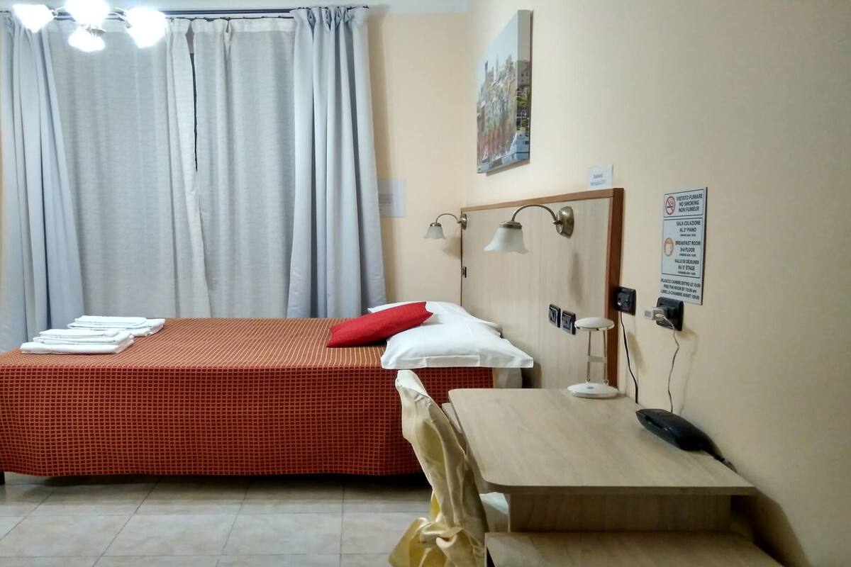 A hotel room with a bed, a desk, a chair, a desk lamp, a telephone, and a window with curtains.