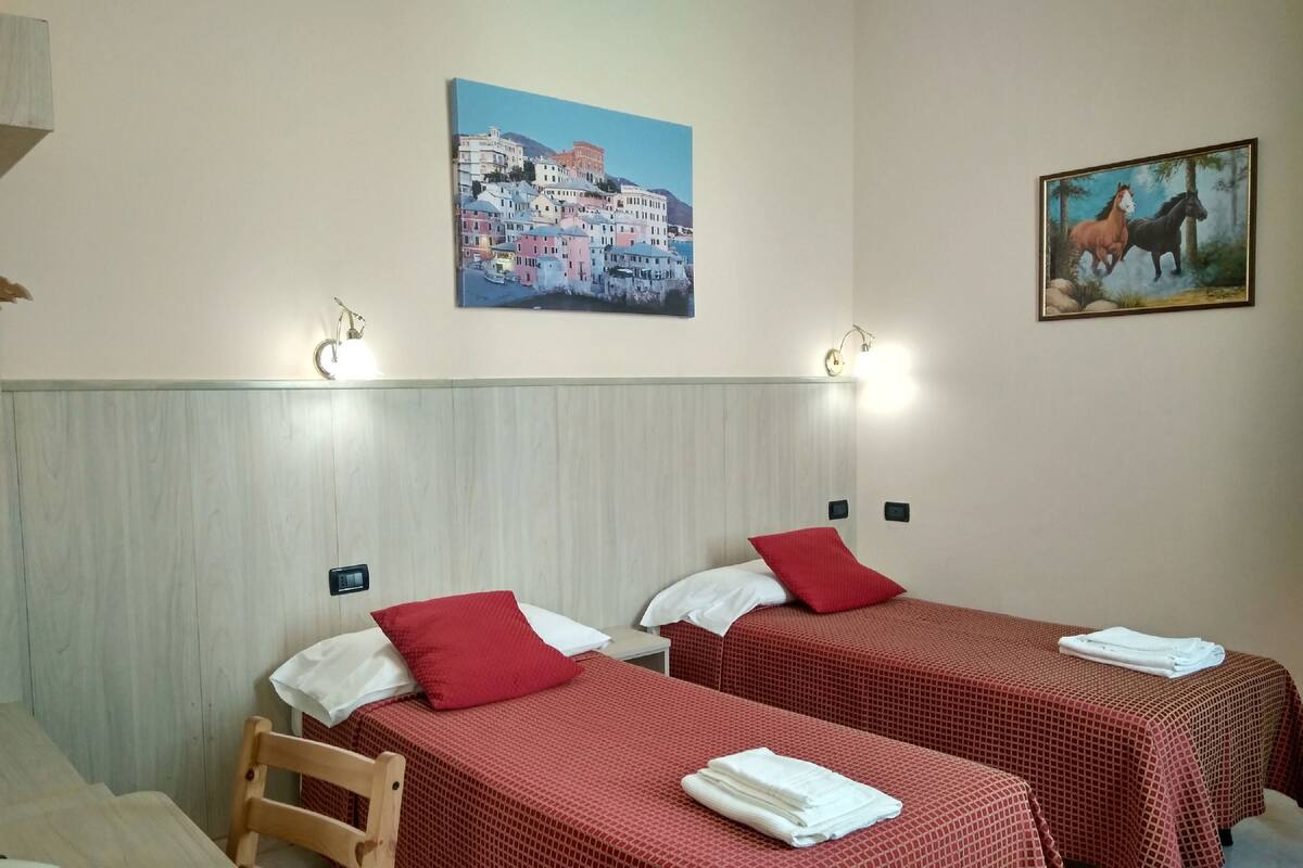 A hotel room with two single beds, each with a red bedspread and white pillows. There is a wooden chair and a small table with a telephone. The room has a wooden headboard and a painting of a coastal town on the wall.