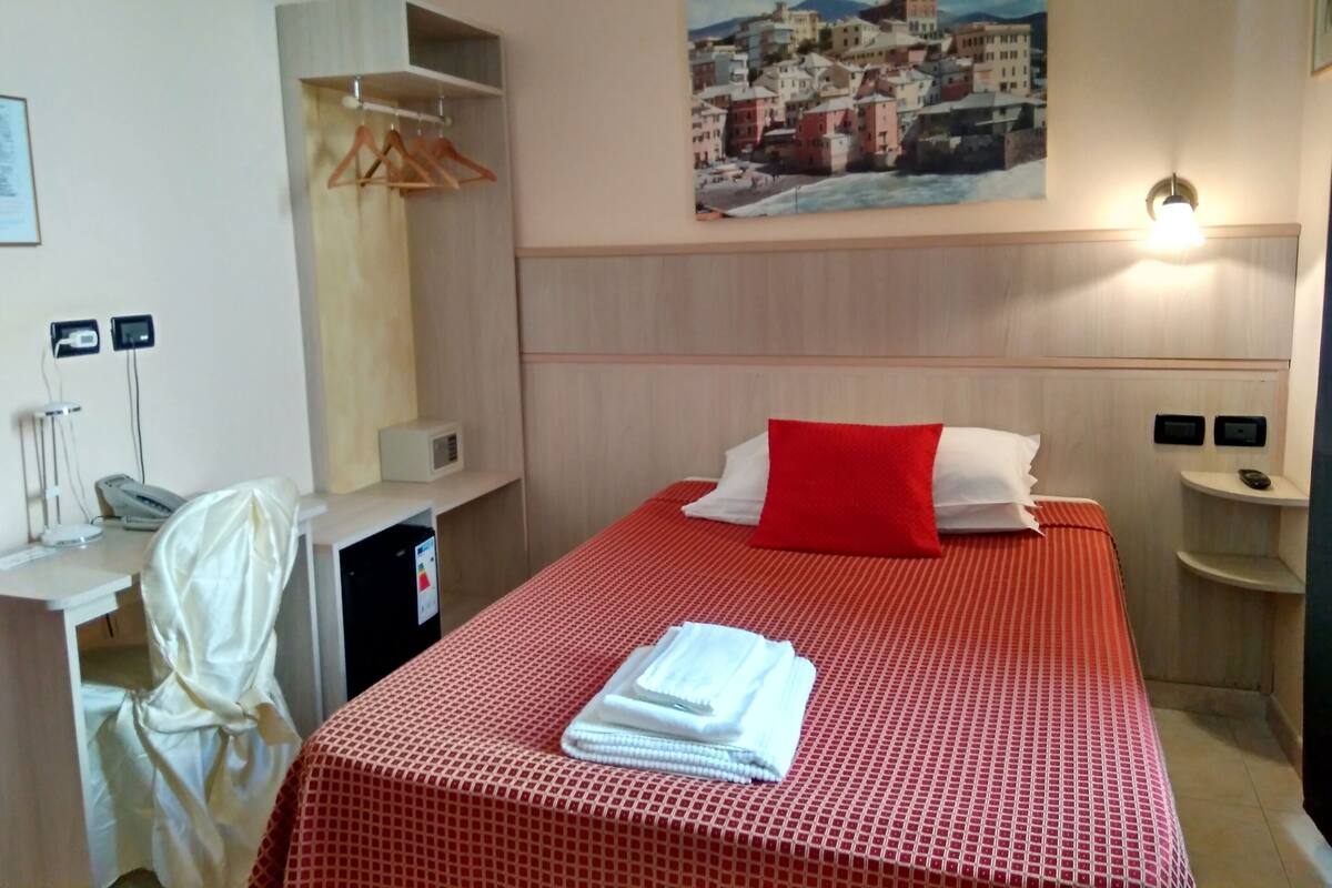 A hotel room with a bed, a painting of a coastal town, a desk with a telephone, and a robe hanging.
