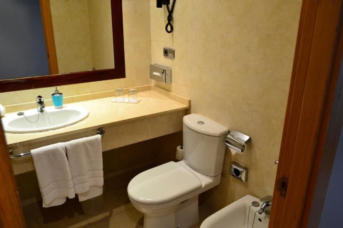 Um banheiro pequeno com vaso sanitário branco, pia e espelho. Há um porta-toalhas com duas toalhas penduradas, um dispensador de sabonete e um suporte para papel higiênico.