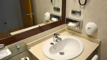 Un baño con lavabo, espejo y diversos servicios como secador de manos, dispensador de toallas de papel y papel higiénico.
