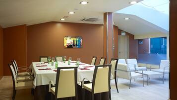 A dining area with a long table set for six, surrounded by chairs. There is a large window providing natural light, and a colorful artwork is visible on the wall.