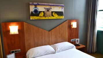 Een hotelkamer met een houten hoofdeinde, twee bedlampjes, een bed met witte lakens, een schilderij boven het hoofdeinde, en een bordje op het nachtkastje.