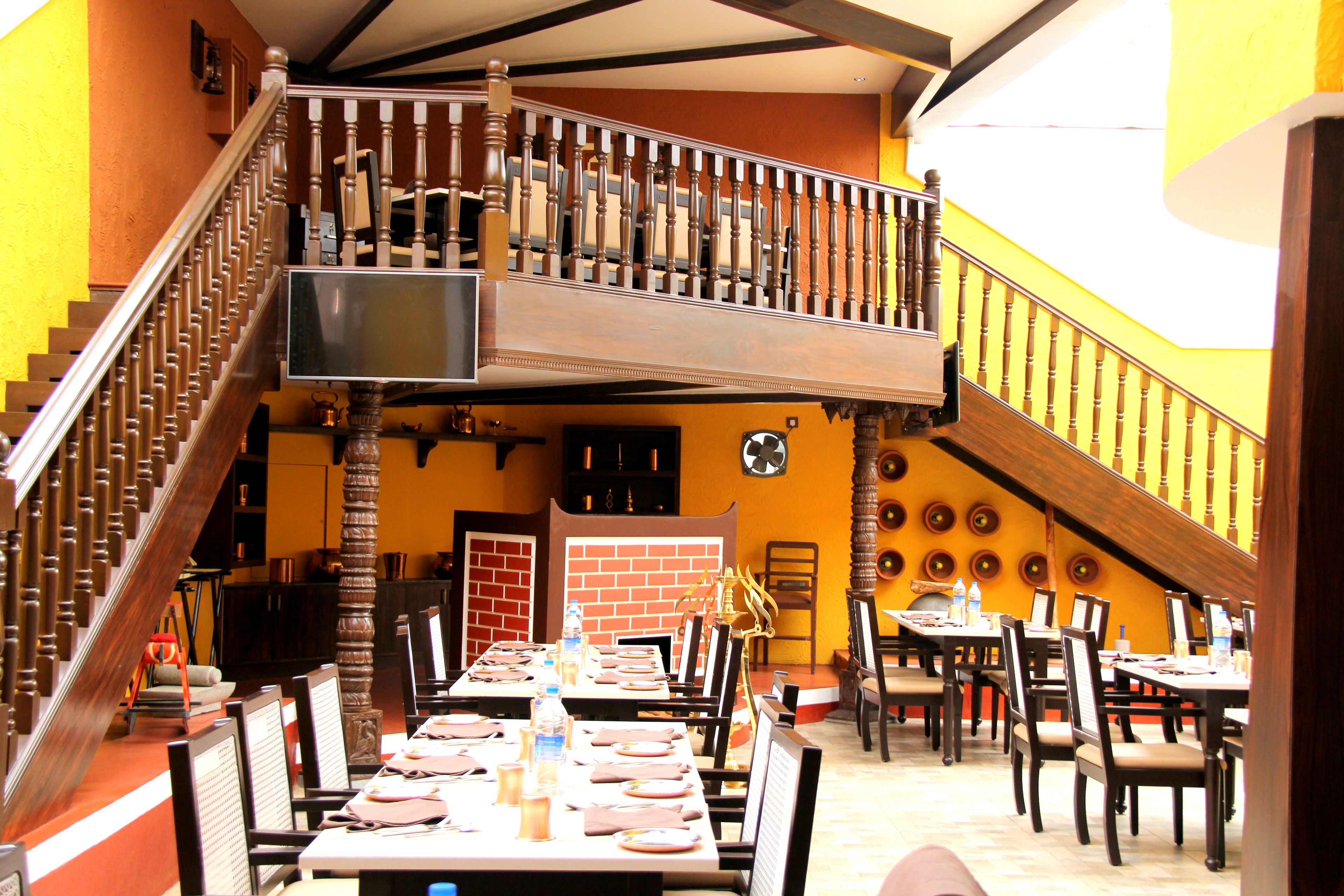 A dining area with tables set for multiple guests, a staircase with wooden balusters, and a brick wall in the background.