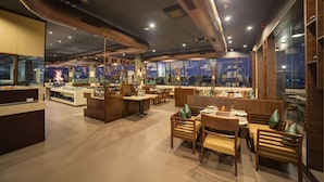 A modern restaurant with wooden furniture and a view of the cityscape through large windows. - Goldfinch Hotel Bangalore (Bengaluru)