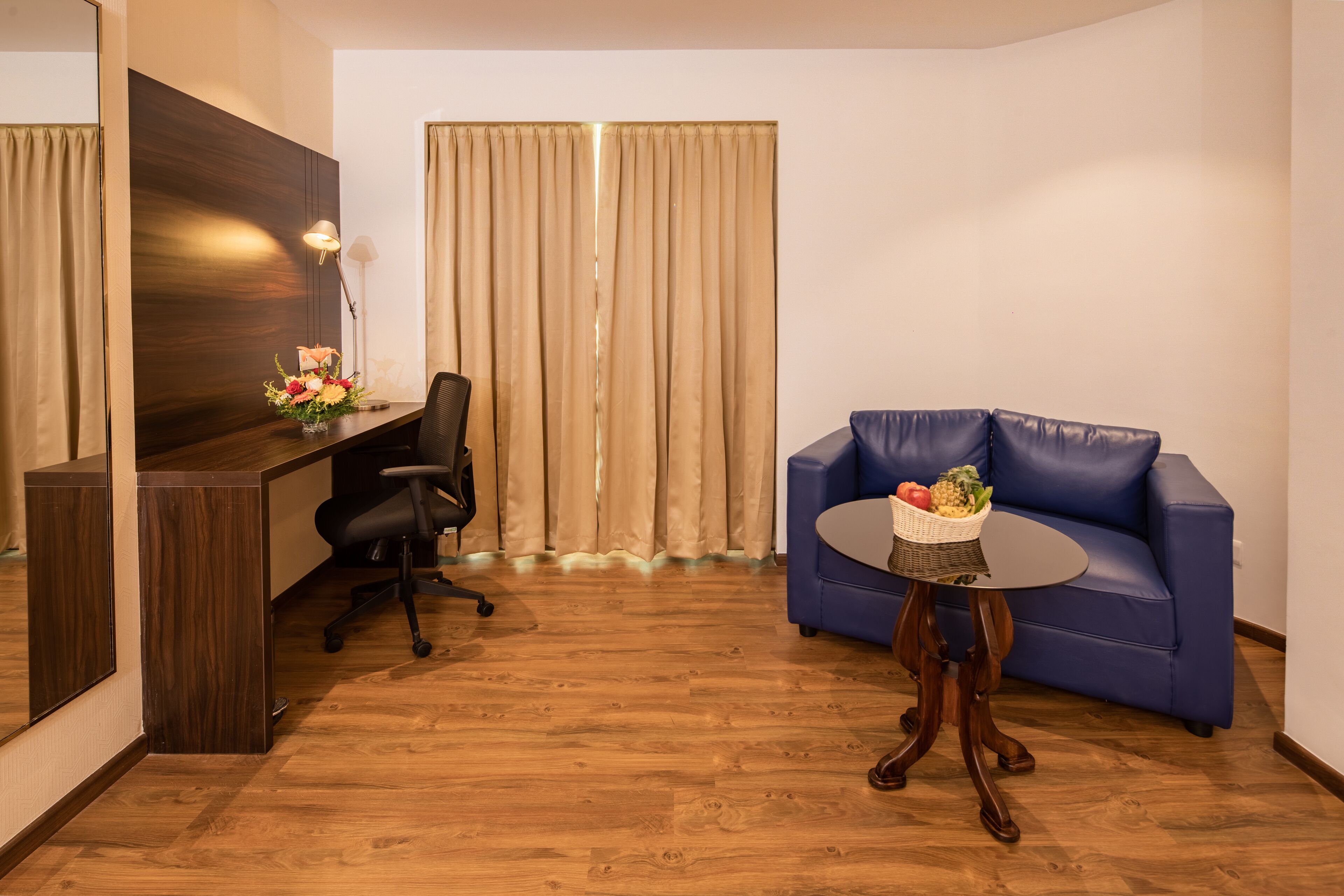 A room with a wooden desk, a blue sofa, a small round table, and a large window with curtains.