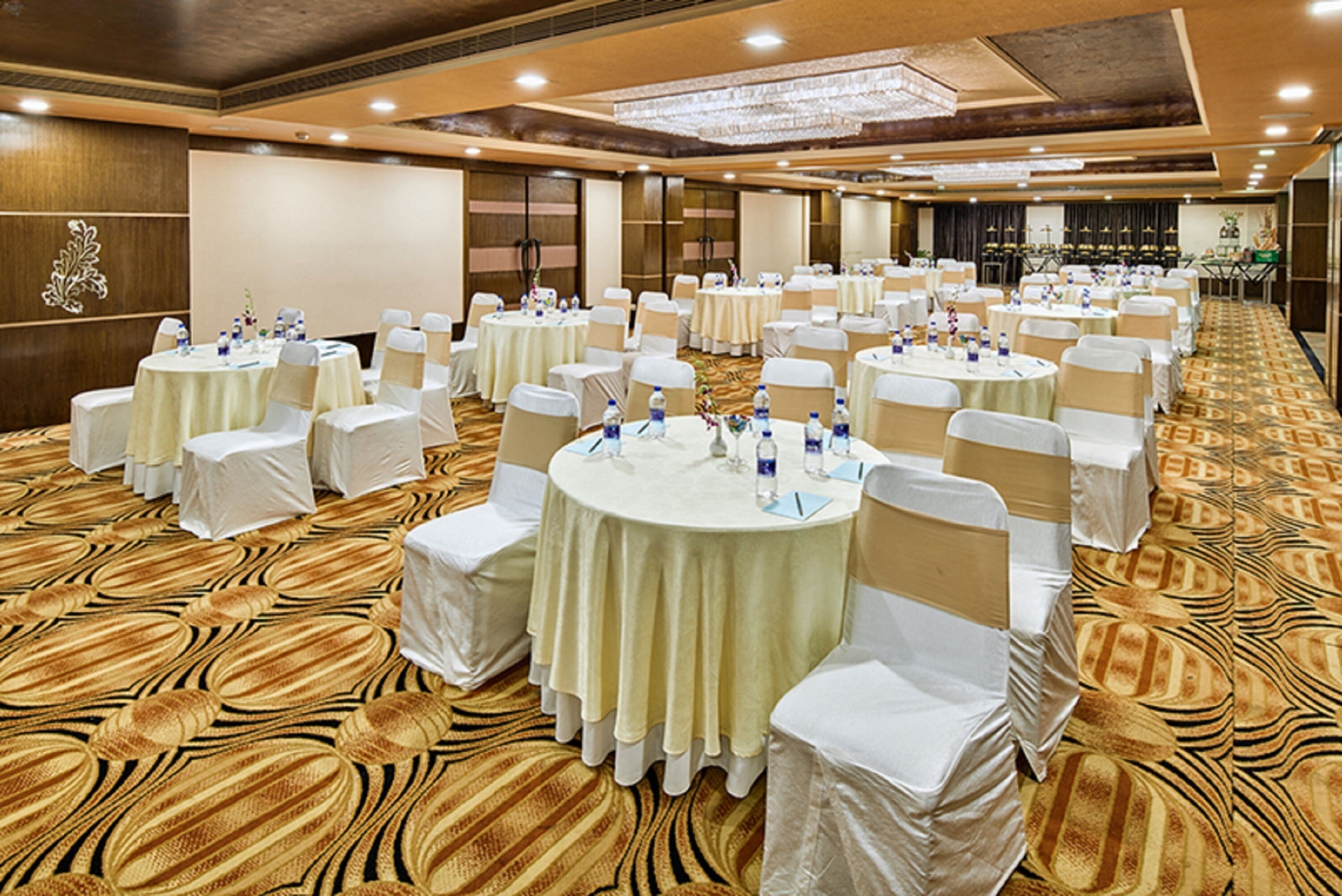 Un amplio salón de banquetes con varias mesas redondas cubiertas con manteles blancos, cada una rodeada de sillas blancas. La sala presenta una alfombra estampada y paneles de madera en las paredes.