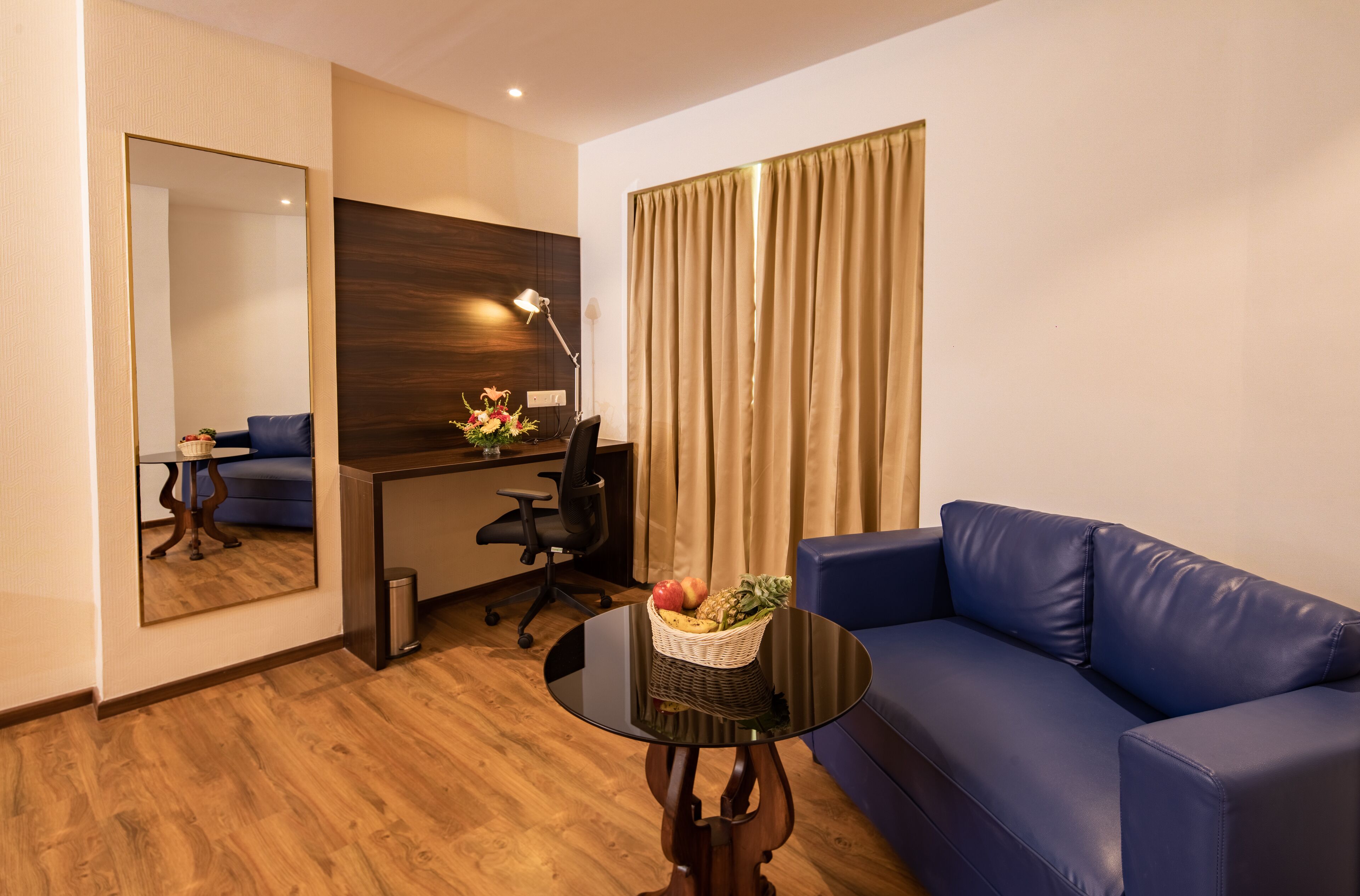 Habitación de hotel con un sofá azul, una mesa redonda negra con una cesta de frutas, un escritorio con una computadora, una silla, un espejo y un ventanal grande con cortinas.