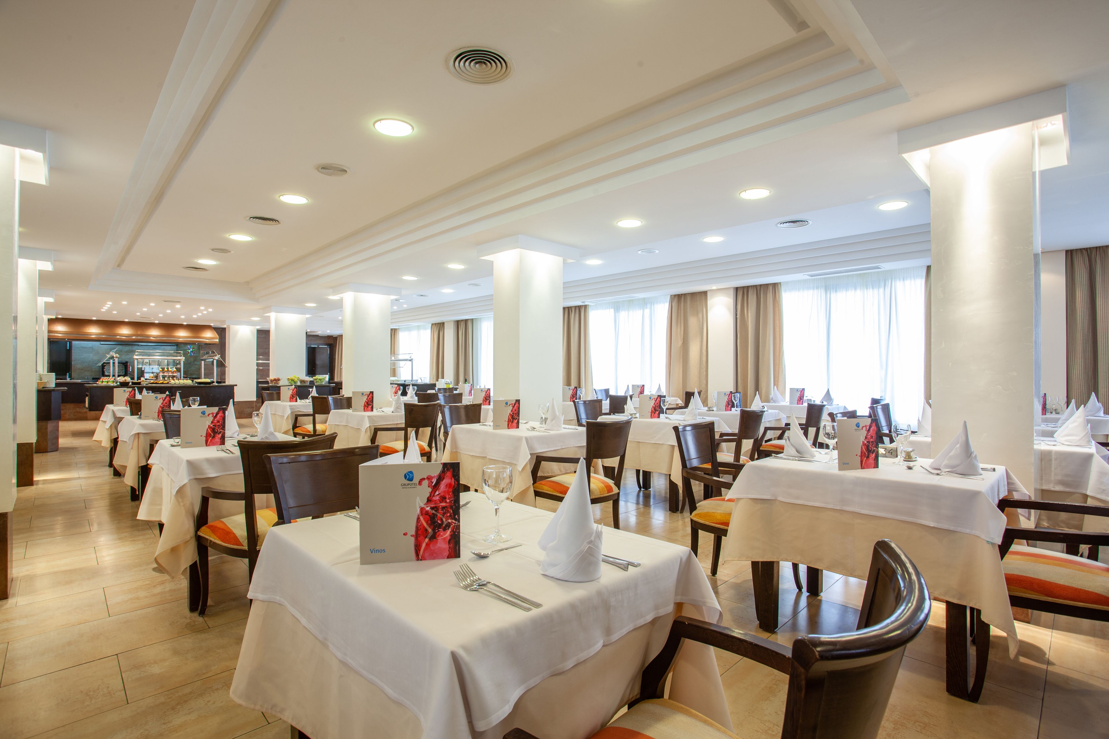 En rummelig hotelrestaurant med flere borde dækket til gæster, med et moderne design, hvide duge og træstole.