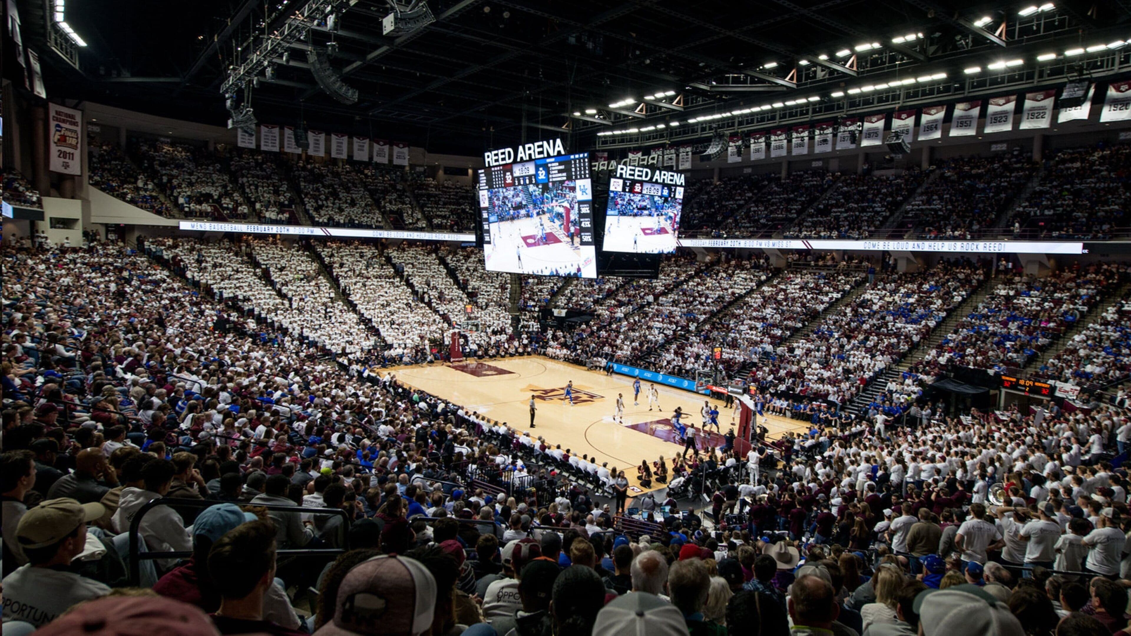 A large indoor arena with multiple tiers of seating, digital scoreboards, and a basketball court.