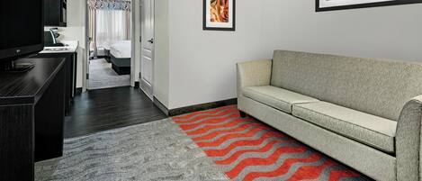 A hotel room with a television, a sofa, a bed, and a hallway leading to another room.