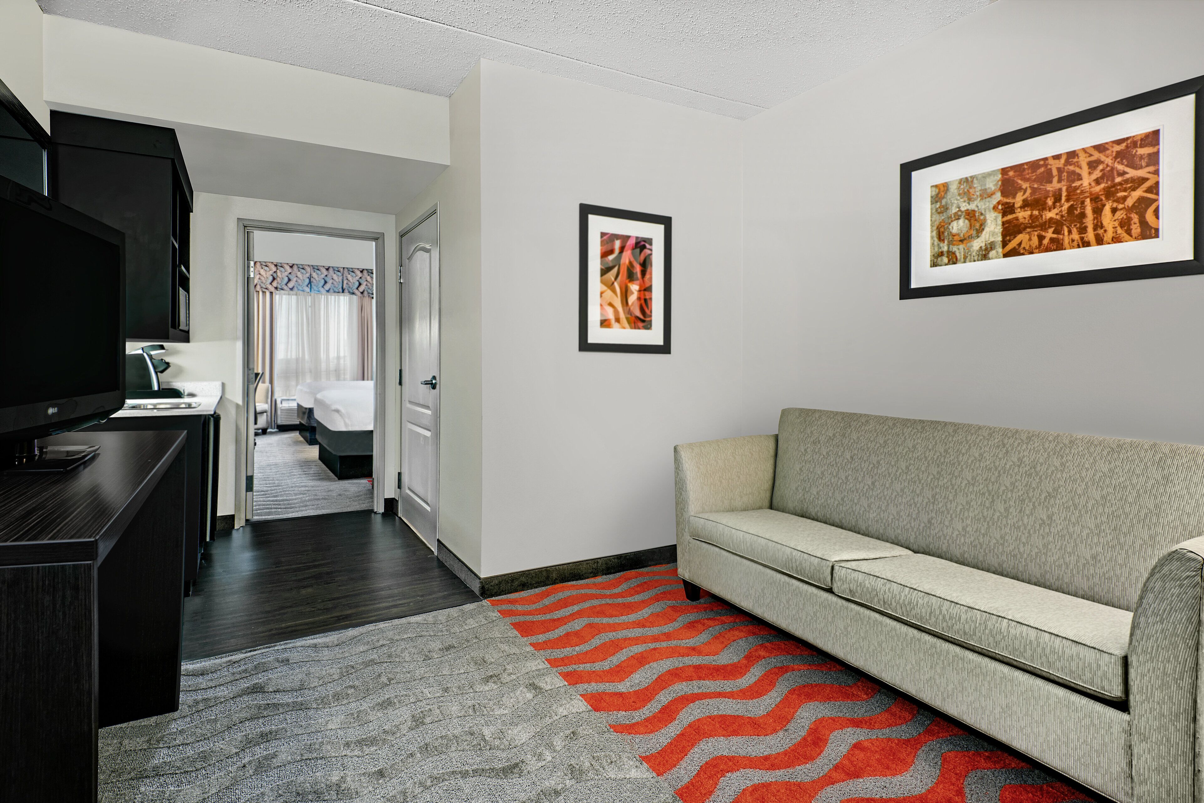 A modern hotel room with a grey sofa, a TV, a desk, and a red and grey rug.