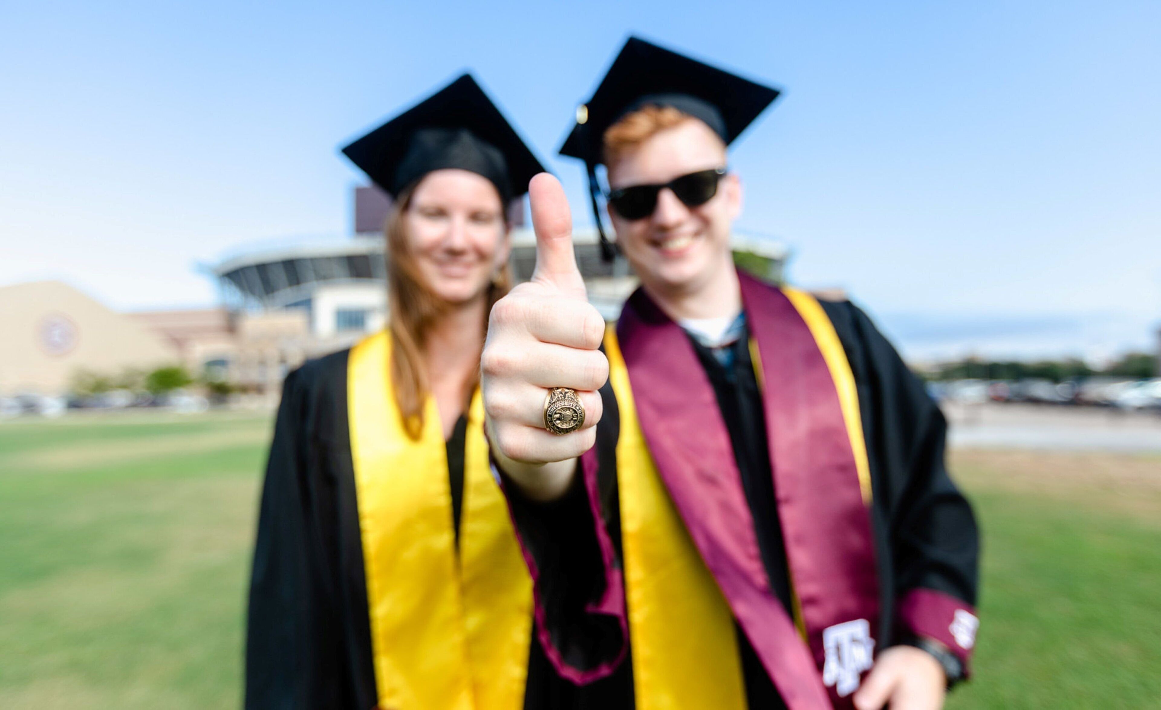 Two graduates in academic gowns and caps, one giving a thumbs-up, standing on a grassy field with a building in the background.