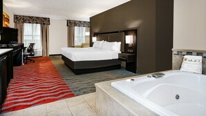 A hotel room with a large bed, a desk with a chair, a TV, a bathroom with a bathtub, and a red and gray striped carpet.