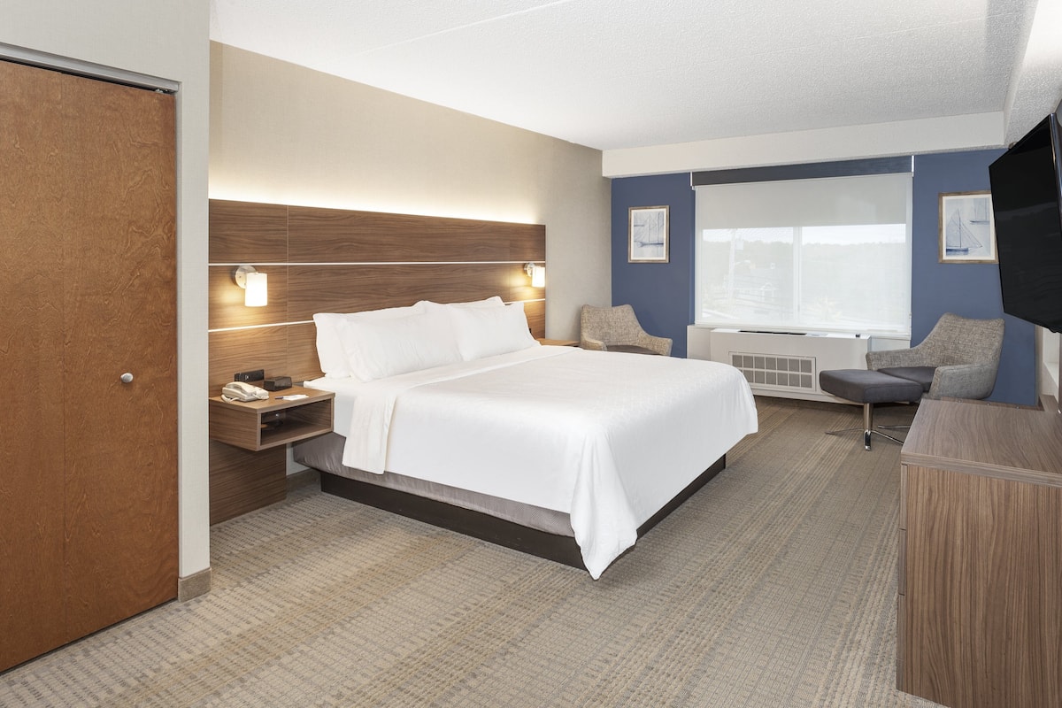A hotel room with a large bed, a wooden headboard, a nightstand, a desk, a chair, and a television.