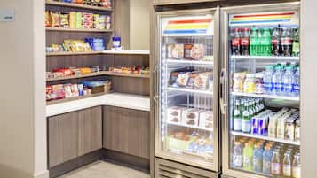 A self-service snack and beverage area with shelves stocked with various snacks and a refrigerated section with bottled water and juices.