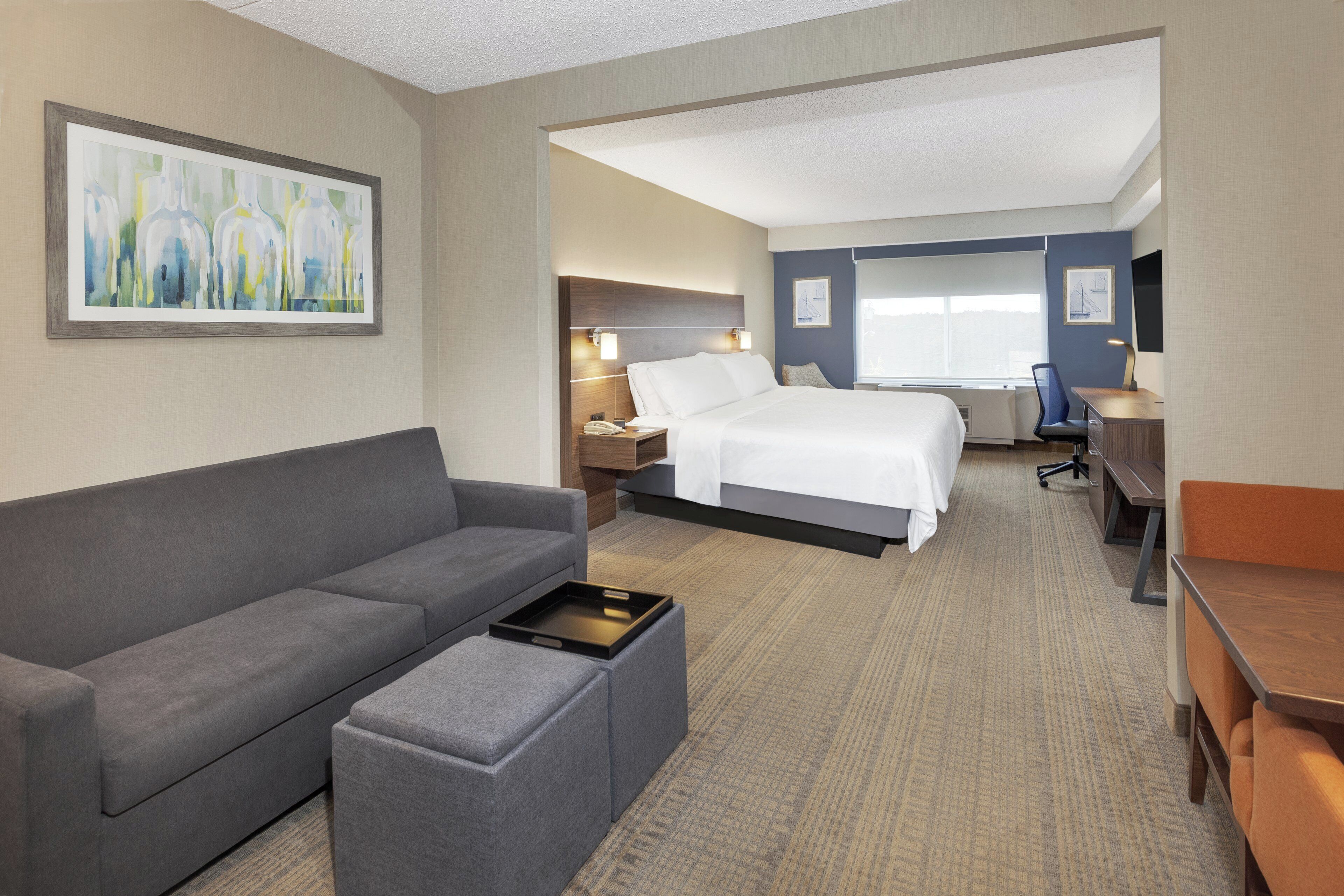 A hotel room with a gray sofa, a small ottoman, a bed with white linens, a desk with a chair, and artwork on the walls.