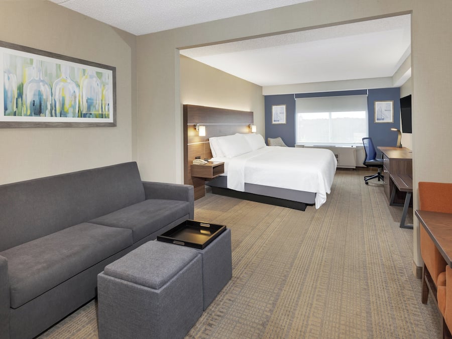 Ein Hotelzimmer mit grauem Sofa, einem kleinen Hocker, einem Bett mit weißer Bettwäsche, einem Schreibtisch mit Stuhl und Kunstwerken an den Wänden.