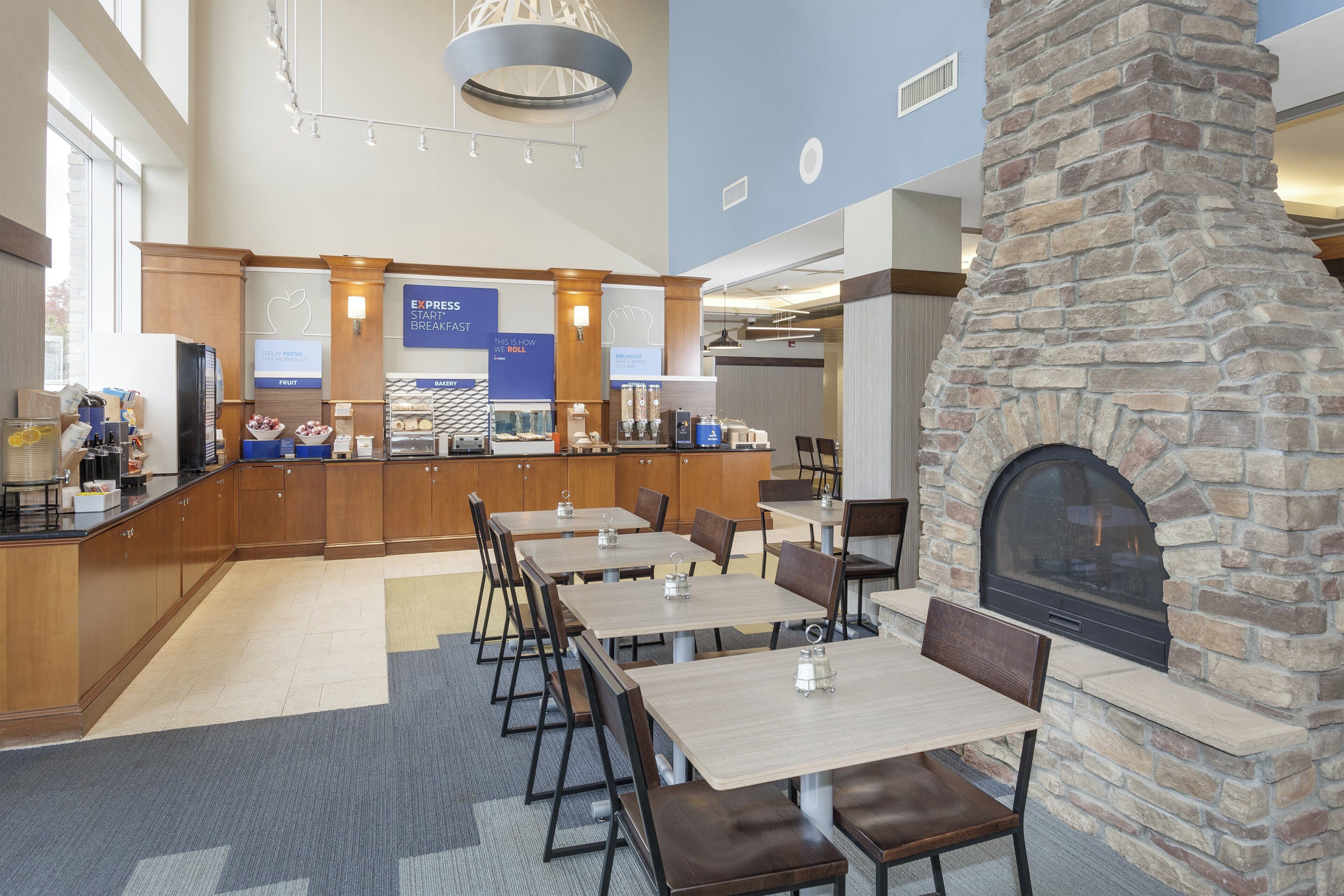 A hotel breakfast area with a stone fireplace, wooden tables, and a fruit display.