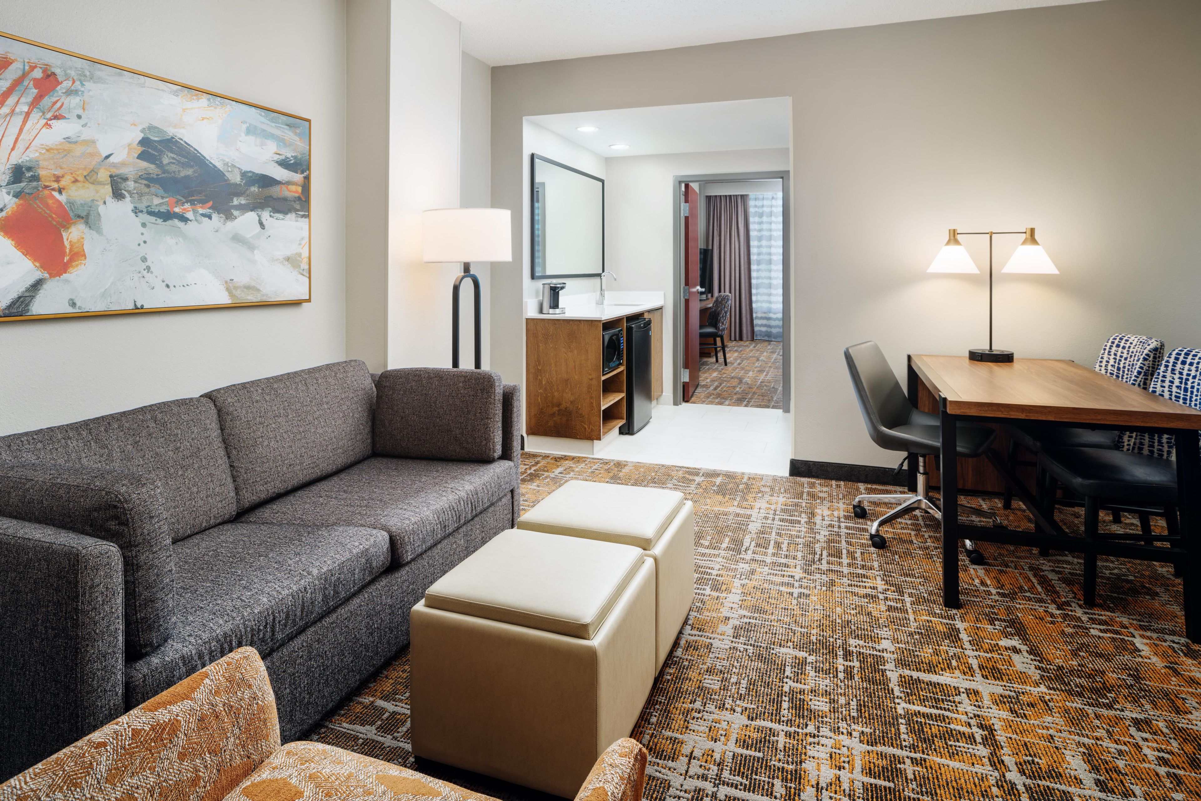 Quarto de hotel moderno com sofá cinzento, um banquinho bege, tapete estampado, mesa de jantar com cadeiras, luminária e um quadro abstrato grande na parede.